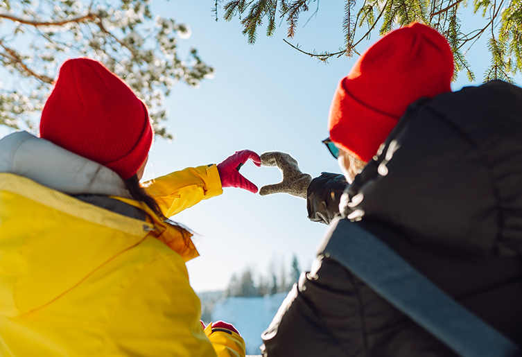 Two people sitting together outside in winter gear with their hands together making a heart