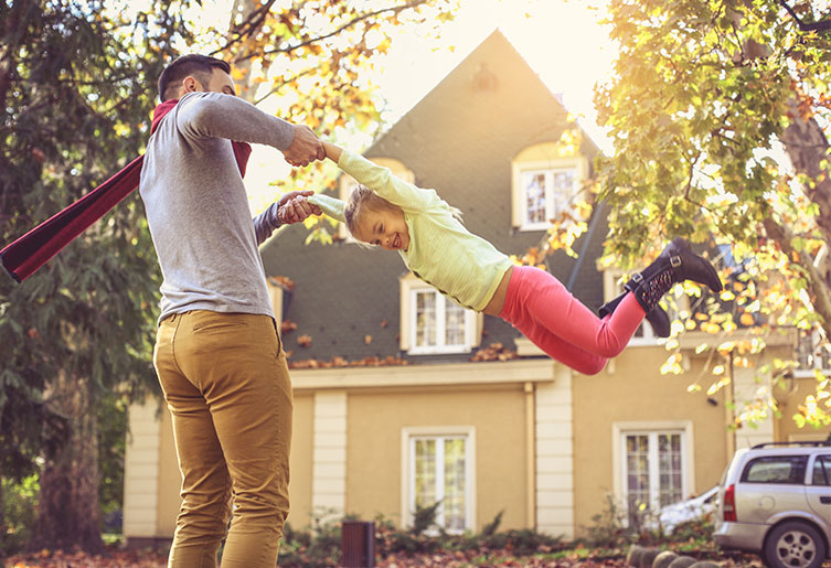 A man swinging his daughter outside their house on a sunny fall day.